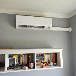 Moja-klima.pl - Biały klimatyzator Rotenso zamontowany na szarej ścianie nad półką z książkami w pokoju. Widoczne białe rury instalacyjne.