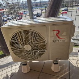 Moja-klima.pl - Klimatyzator Rotenso na balkonie z widokiem na parking. Jednostka zewnętrzna z logo i nazwą marki, zamontowana na specjalnych nóżkach. Widoczne elementy instalacji.