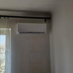 Moja-klima.pl - Biały klimatyzator ścienny z wyświetlaczem temperatury (16 stopni) zamontowany w pokoju z oknem zasłoniętym firanką. Widok z dołu.