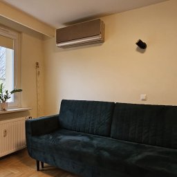 Moja-klima.pl - Klimatyzator w salonie z zieloną sofą i rośliną w oknie. Widok na balkon. Beżowe ściany, drewniana podłoga. Nowoczesny design, neutralne kolory.