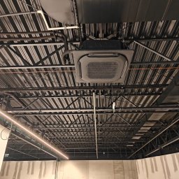 Moja-klima.pl - Sufit biurowy z zamontowaną kasetonową jednostką klimatyzacji, widoczne rury i instalacje pod stropem. Oświetlenie liniowe LED. Surowy beton na ścianach.