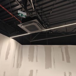Moja-klima.pl - Czarna, sufitowa klimatyzacja kasetonowa w surowym wnętrzu biurowym z widoczną instalacją i oświetleniem LED. Ściany w trakcie przygotowania do malowania.