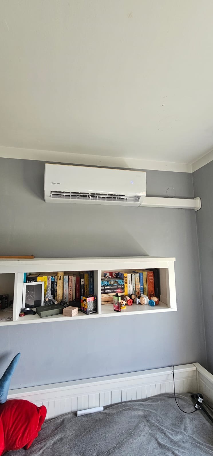Biały klimatyzator Rotenso zamontowany na szarej ścianie nad półką z książkami w pokoju. Widoczne białe rury instalacyjne.