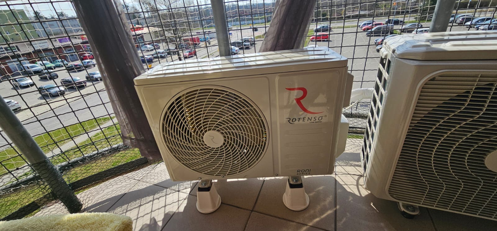 Klimatyzator Rotenso na balkonie z widokiem na parking. Jednostka zewnętrzna z logo i nazwą marki, zamontowana na specjalnych nóżkach. Widoczne elementy instalacji.