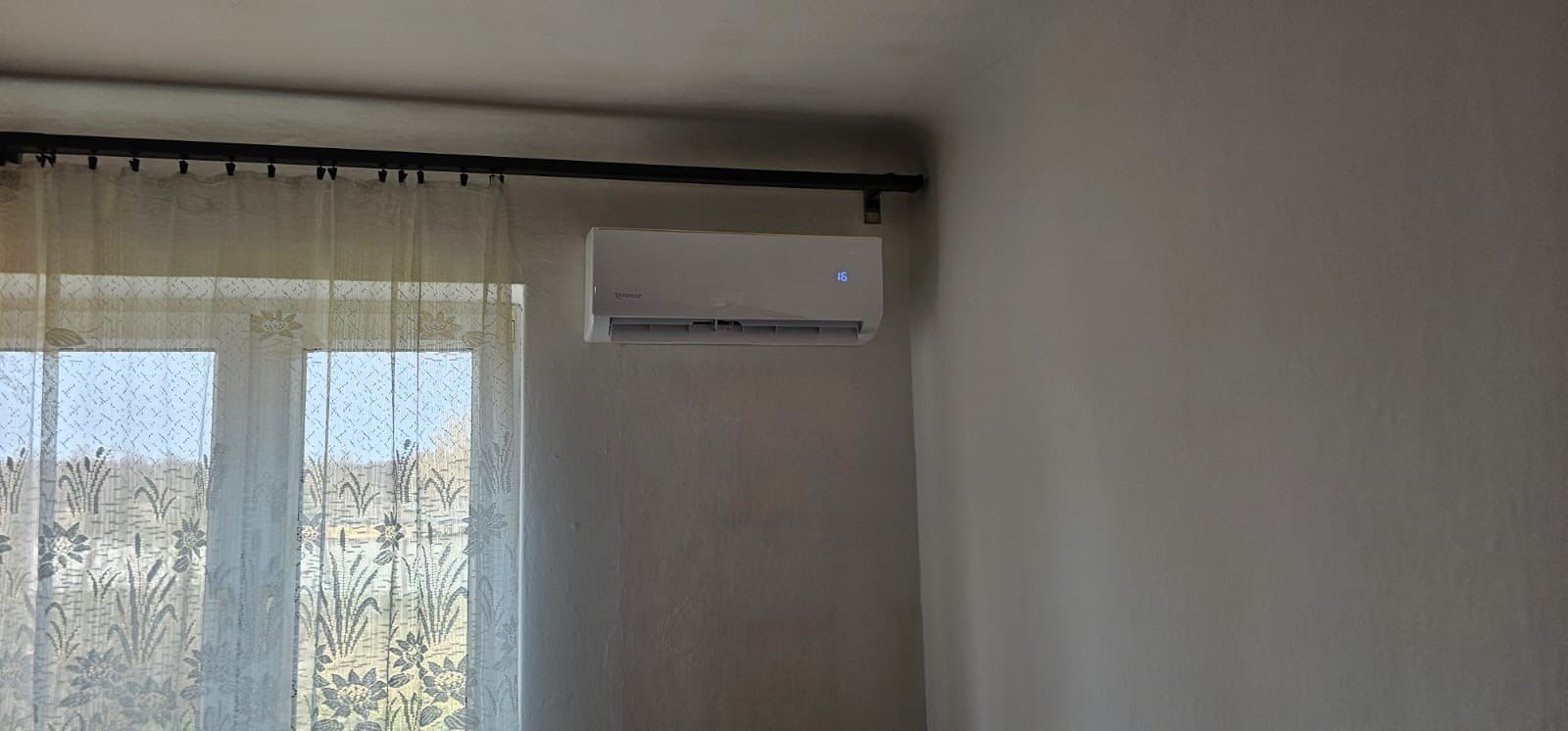 Biały klimatyzator ścienny z wyświetlaczem temperatury (16 stopni) zamontowany w pokoju z oknem zasłoniętym firanką. Widok z dołu.