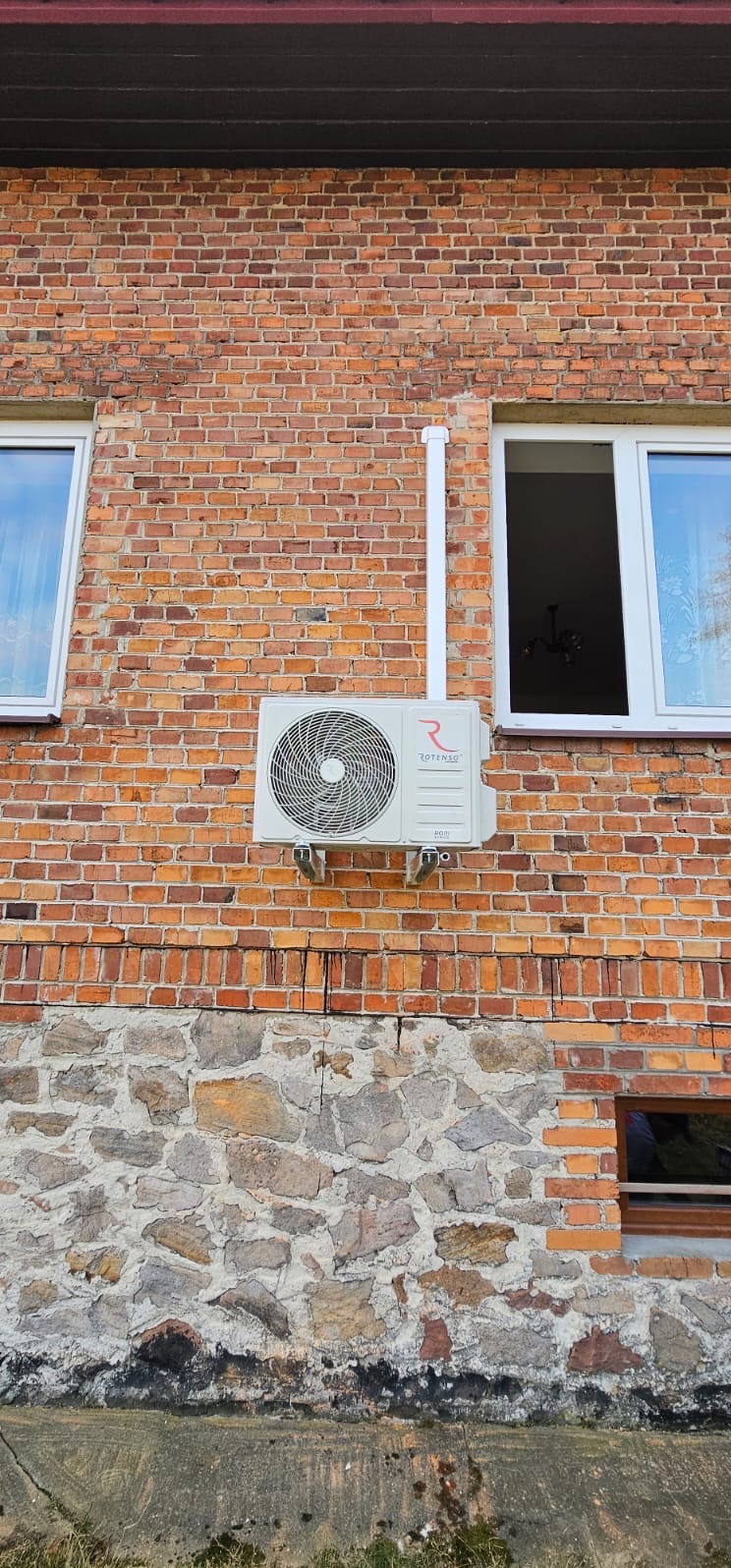 Biały klimatyzator Rotenso zamontowany na ceglanej ścianie budynku z kamienną podmurówką, widoczny biały przewód odprowadzający skropliny i okno z białymi ramami.