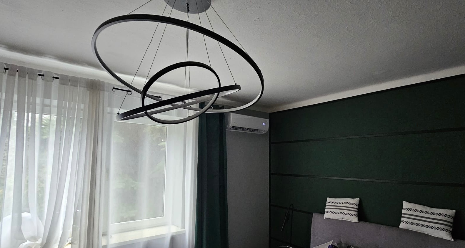 Nowoczesny klimatyzator ścienny w jasnym pokoju z designerską lampą. Widoczne białe zasłony i ciemnozielona ściana z dekoracyjnymi listwami.