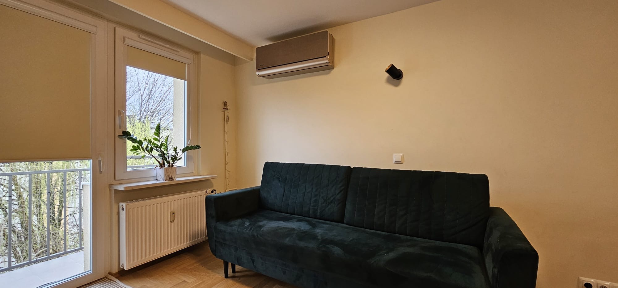 Klimatyzator w salonie z zieloną sofą i rośliną w oknie. Widok na balkon. Beżowe ściany, drewniana podłoga. Nowoczesny design, neutralne kolory.
