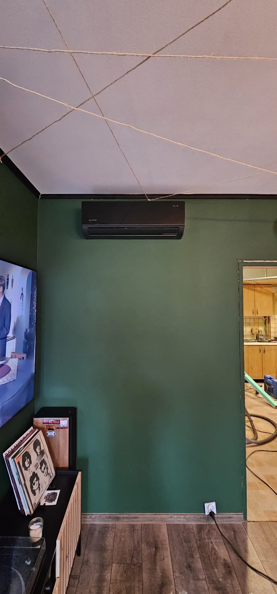 Czarny klimatyzator na zielonej ścianie w pokoju z elementami dekoracyjnymi z sznurka na suficie. Widoczny fragment kuchni w tle.