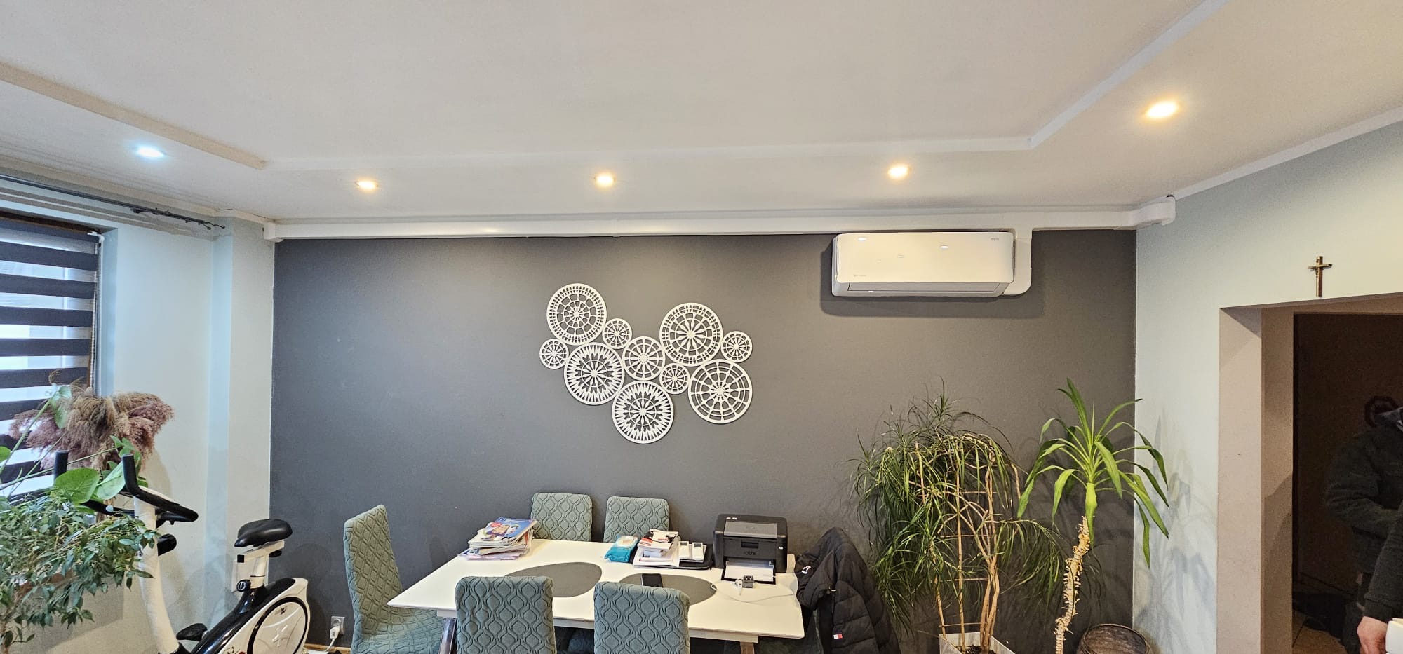 Biały klimatyzator ścienny zamontowany w pokoju nad dekoracją z ażurowych kół. Widoczne rośliny doniczkowe, stół z krzesłami i sprzęt do ćwiczeń.