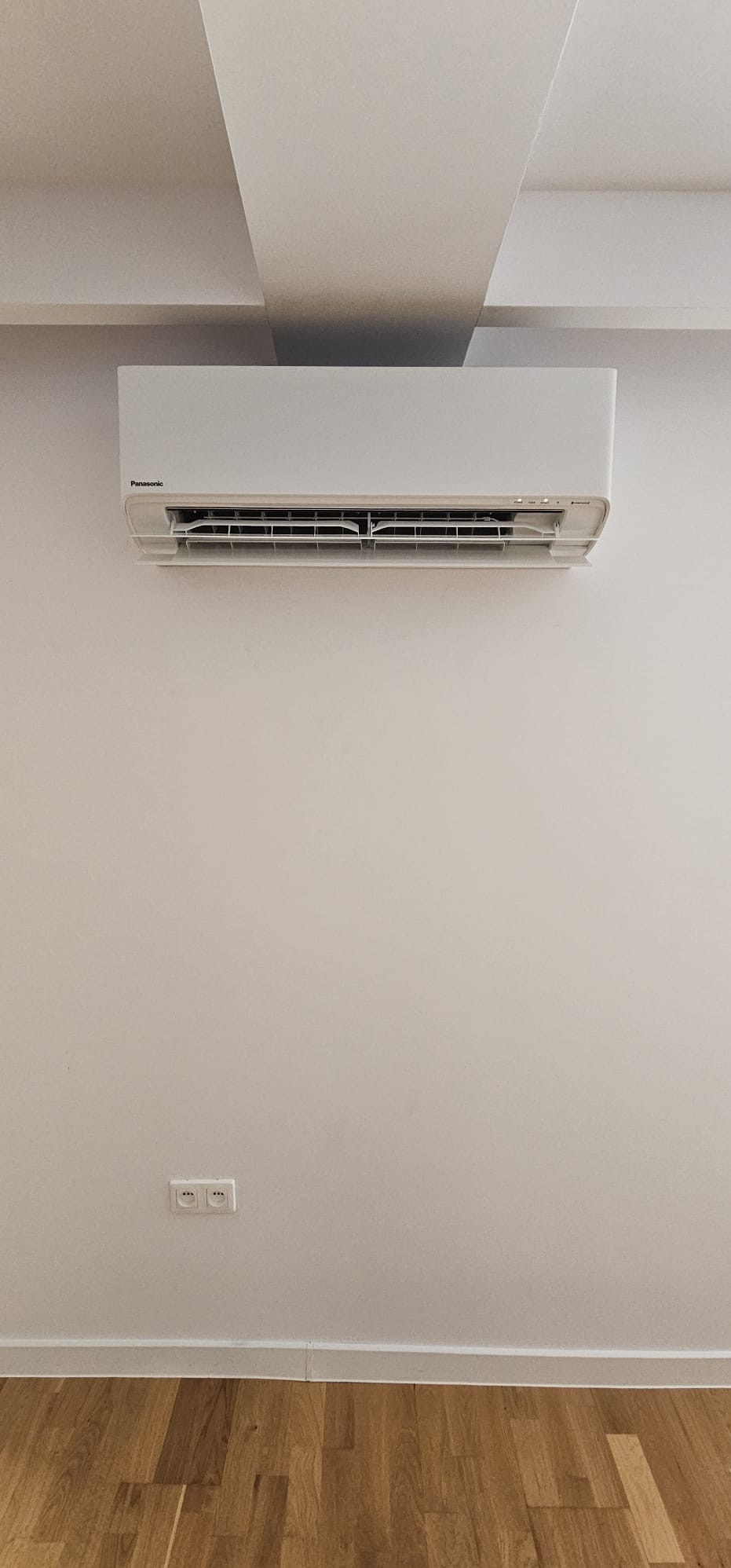 Biały klimatyzator Panasonic zamontowany na ścianie w jasnym pomieszczeniu z drewnianą podłogą. Widoczne gniazdka elektryczne pod klimatyzatorem.