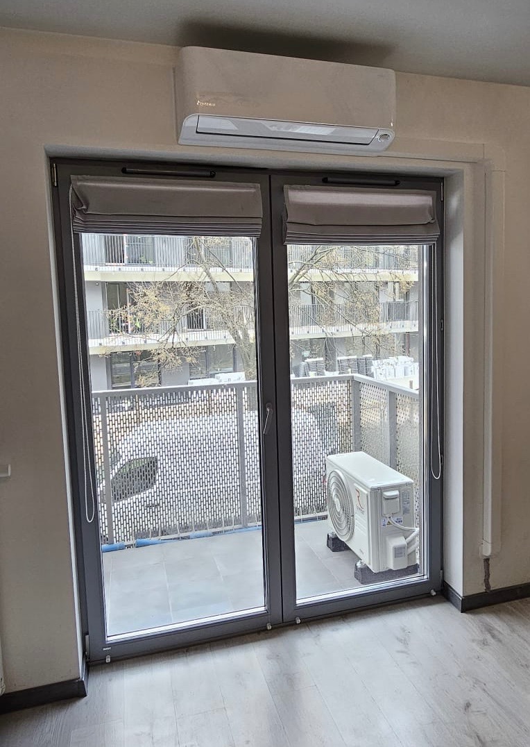 Biały klimatyzator na ścianie nad oknem balkonowym z szarymi ramami i roletami rzymskimi. Na balkonie widoczna jednostka zewnętrzna klimatyzacji.
