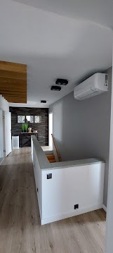 Biała jednostka klimatyzacyjna zamontowana na szarej ścianie w nowoczesnym wnętrzu domu z drewnianymi akcentami i schodami. Widok z dołu.