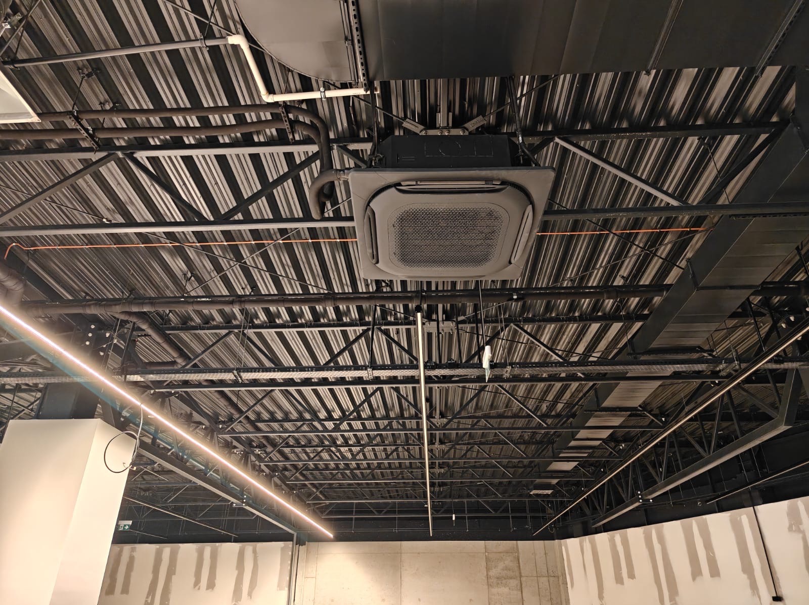 Sufit biurowy z zamontowaną kasetonową jednostką klimatyzacji, widoczne rury i instalacje pod stropem. Oświetlenie liniowe LED. Surowy beton na ścianach.