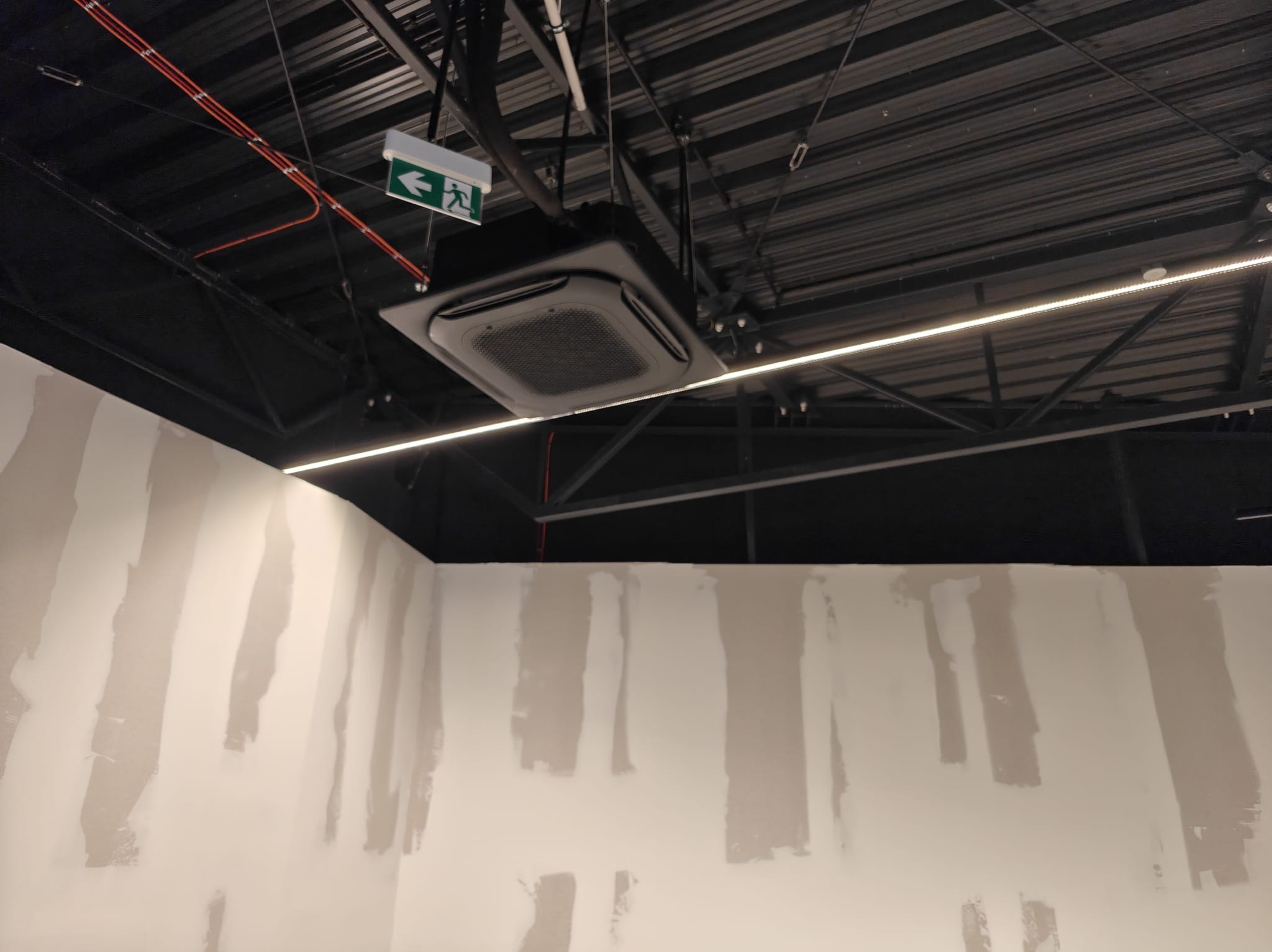 Czarna, sufitowa klimatyzacja kasetonowa w surowym wnętrzu biurowym z widoczną instalacją i oświetleniem LED. Ściany w trakcie przygotowania do malowania.