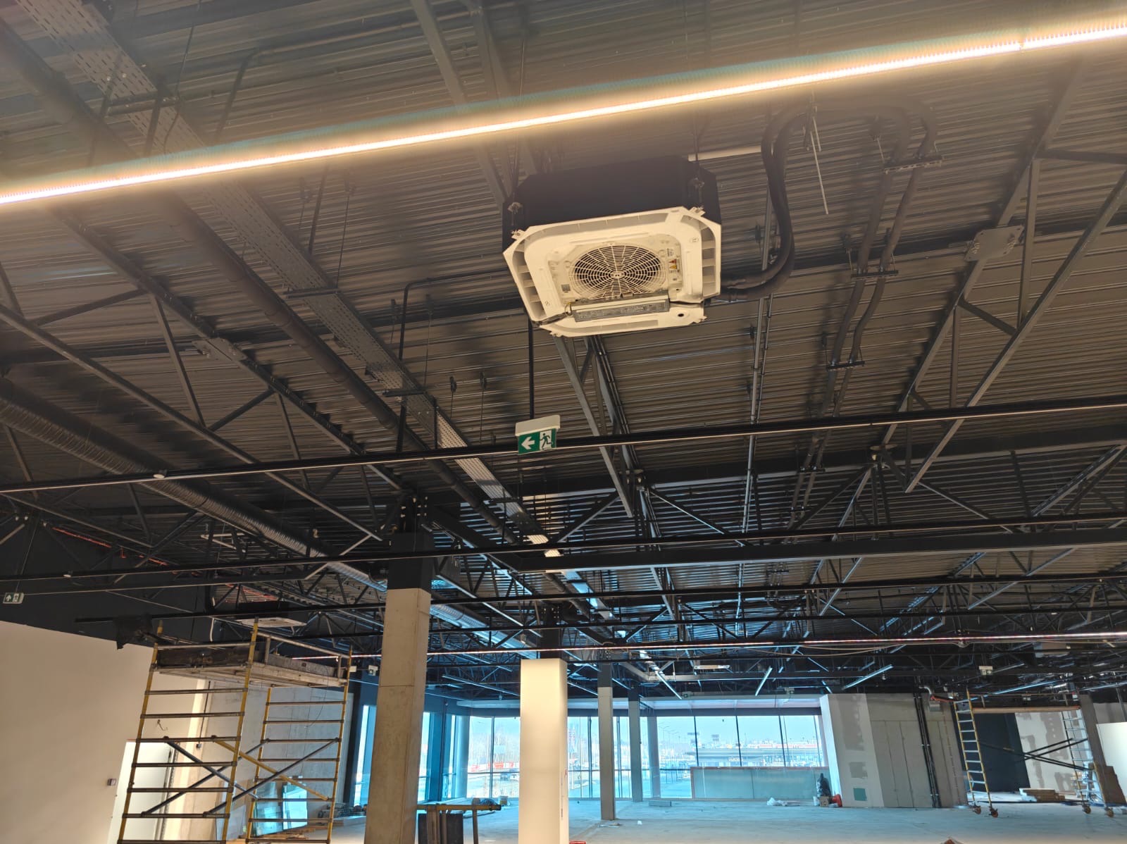 Centralna jednostka klimatyzacji sufitowej w surowym wnętrzu biurowym, widoczne rury i instalacje, rusztowanie w tle, duże okna z widokiem na miasto.