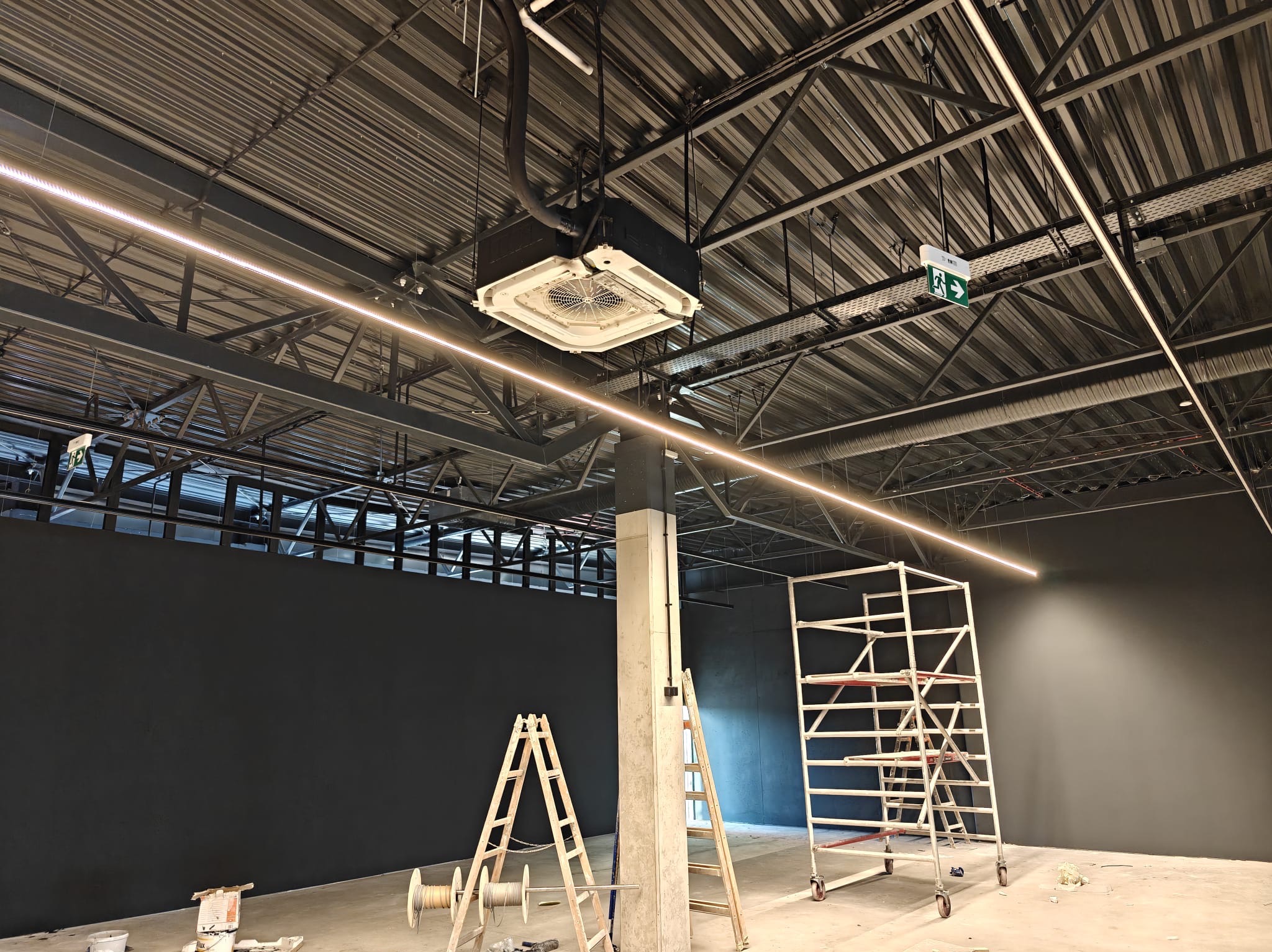 Montaż sufitowej klimatyzacji kasetonowej w hali, widoczne drabiny i rusztowanie, surowy stan wykończenia, oświetlenie liniowe LED.