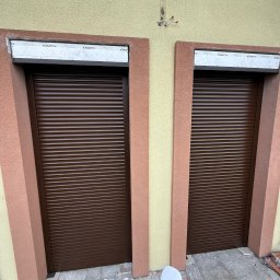 H&I Automatyka Marcin Rozmarynowski - Dwa brązowe okna z zamkniętymi żaluzjami zewnętrznymi na elewacji budynku z beżowo-różowymi detalami, sterowane pilotem trzymanym w dłoni