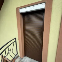 Brązowa roleta zewnętrzna opuszczona na okno, widoczne fragmenty zabezpieczeń montażowych, ściana budynku z dekoracyjnym tynkiem w dwóch odcieniach.