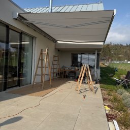 H&I Automatyka Marcin Rozmarynowski - Montaż jasnej, rozkładanej osłony przeciwsłonecznej nad tarasem domu jednorodzinnego, widoczne drabiny, narzędzia i elementy montażowe.