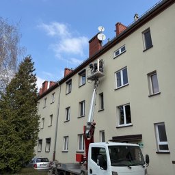 H&I Automatyka Marcin Rozmarynowski - Montażysta w podnośniku koszowym instaluje osłonę okienną na elewacji budynku mieszkalnego, widoczny samochód dostawczy z podnośnikiem i fragment zieleni.