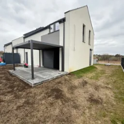 Nowoczesna, szara pergola metalowa z roletą zewnętrzną przy białym domu z kostką brukową na patio, widok z perspektywy ogrodu w pochmurny dzień.