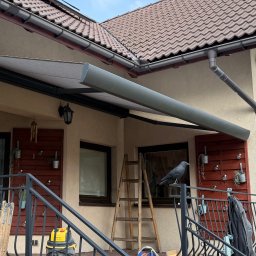 H&I Automatyka Marcin Rozmarynowski - Szara, zwinięta markiza tarasowa zamontowana nad balkonem domu z brązową dachówką, widoczna drabina, dekoracje balkonowe i atrapa kruka na balustradzie.