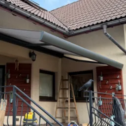 Szara, zwinięta markiza tarasowa zamontowana nad balkonem domu z brązową dachówką, widoczna drabina, dekoracje balkonowe i atrapa kruka na balustradzie.