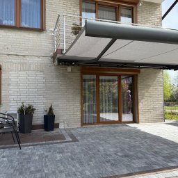 H&I Automatyka Marcin Rozmarynowski - Zewnętrzna pergola metalowa z jasną tkaniną zamontowana nad tarasem przy domu z jasnej cegły, widoczne okna z roletami zewnętrznymi w brązowych ramach oraz szara kostka brukowa.