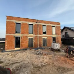 Surowy stan budynku z czerwonej cegły z zamontowanymi oknami i drzwiami, widoczna betoniarka i materiały budowlane na placu budowy.