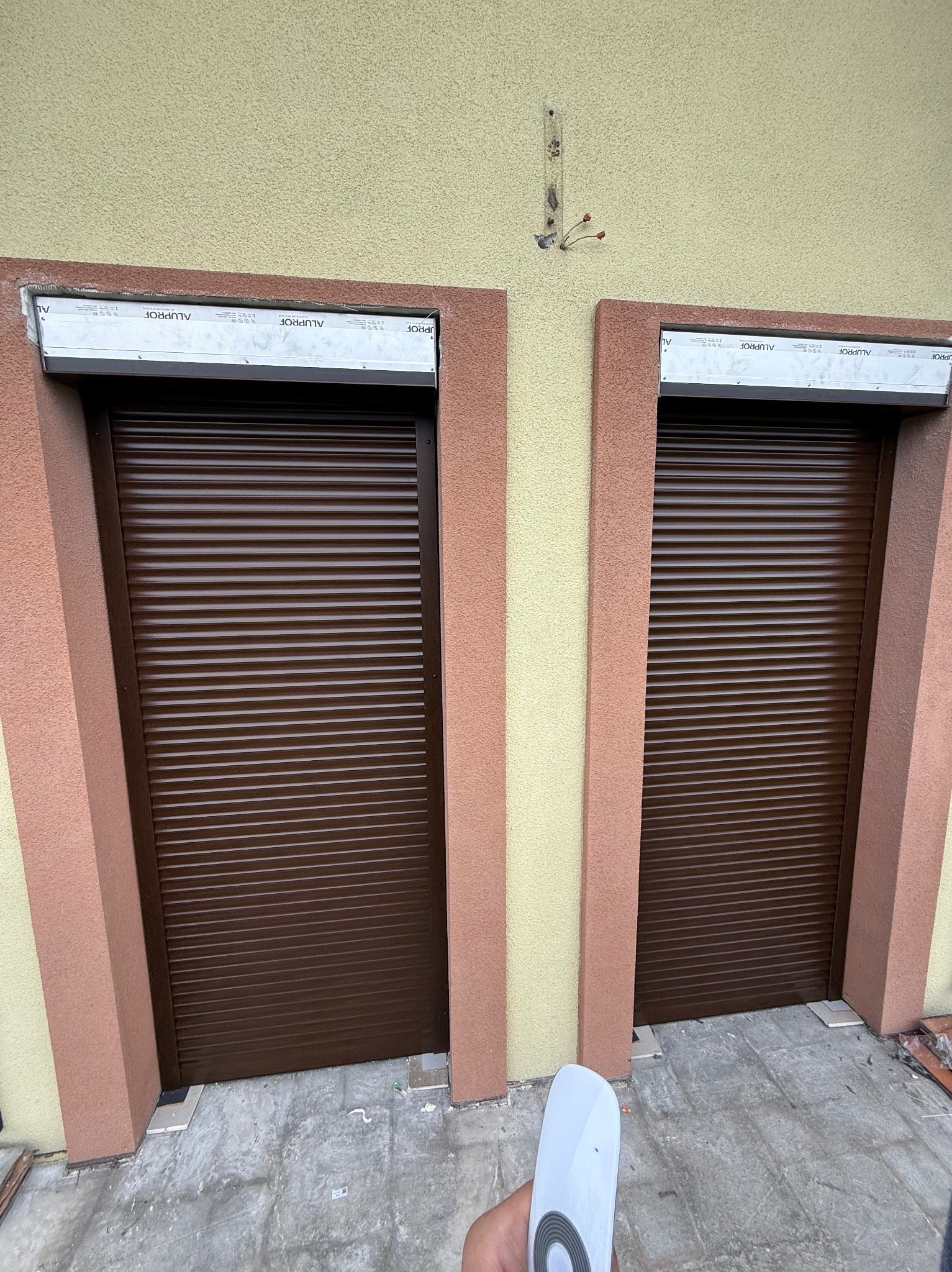 Dwa brązowe okna z zamkniętymi żaluzjami zewnętrznymi na elewacji budynku z beżowo-różowymi detalami, sterowane pilotem trzymanym w dłoni