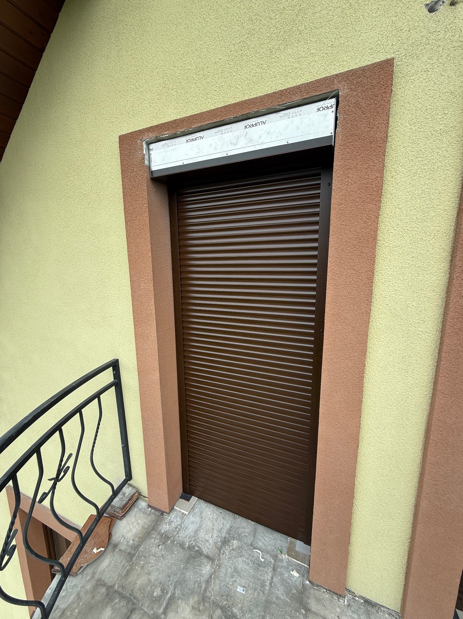 Brązowa roleta zewnętrzna opuszczona na okno, widoczne fragmenty zabezpieczeń montażowych, ściana budynku z dekoracyjnym tynkiem w dwóch odcieniach.