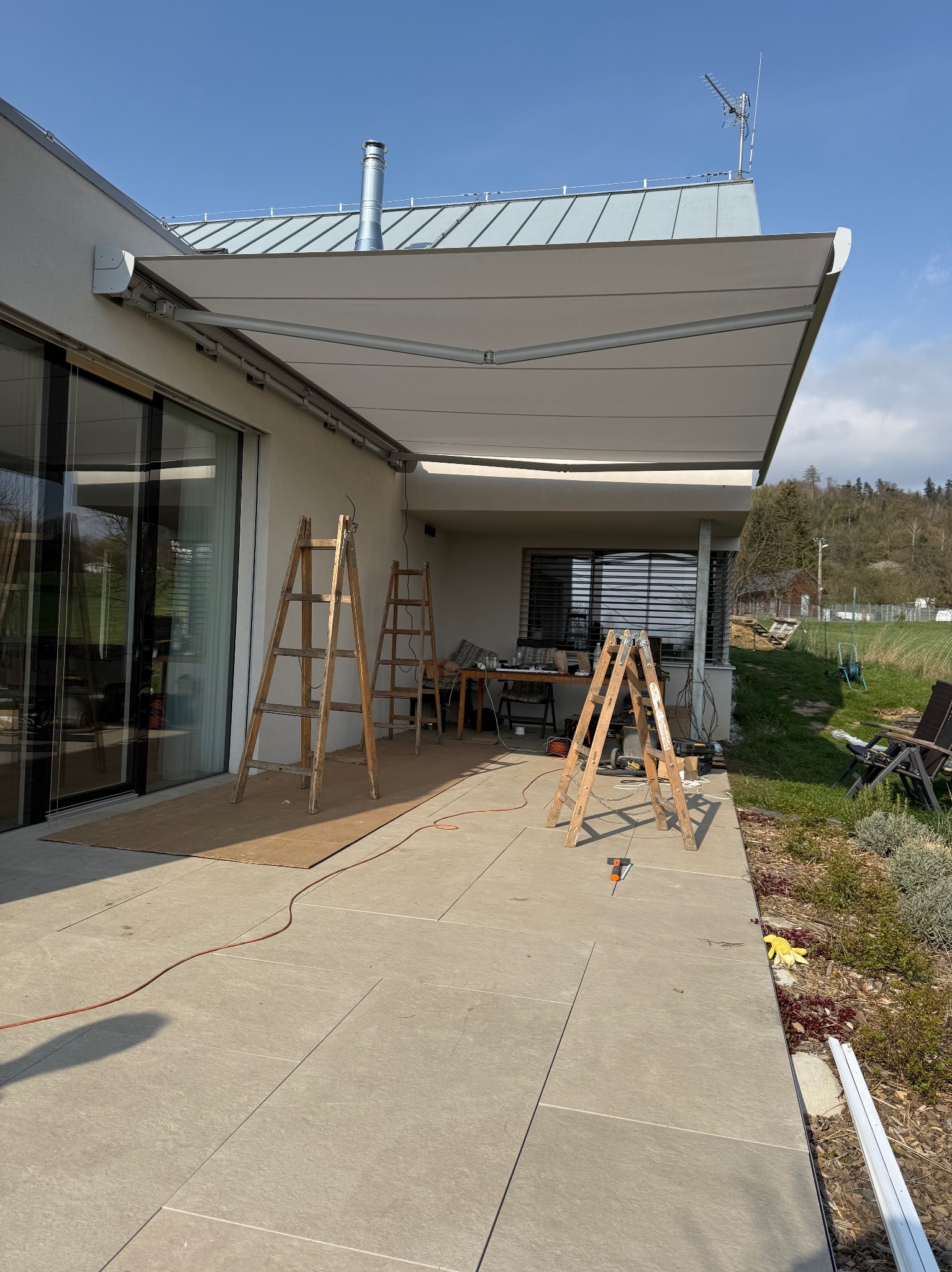 Montaż jasnej, rozkładanej osłony przeciwsłonecznej nad tarasem domu jednorodzinnego, widoczne drabiny, narzędzia i elementy montażowe.