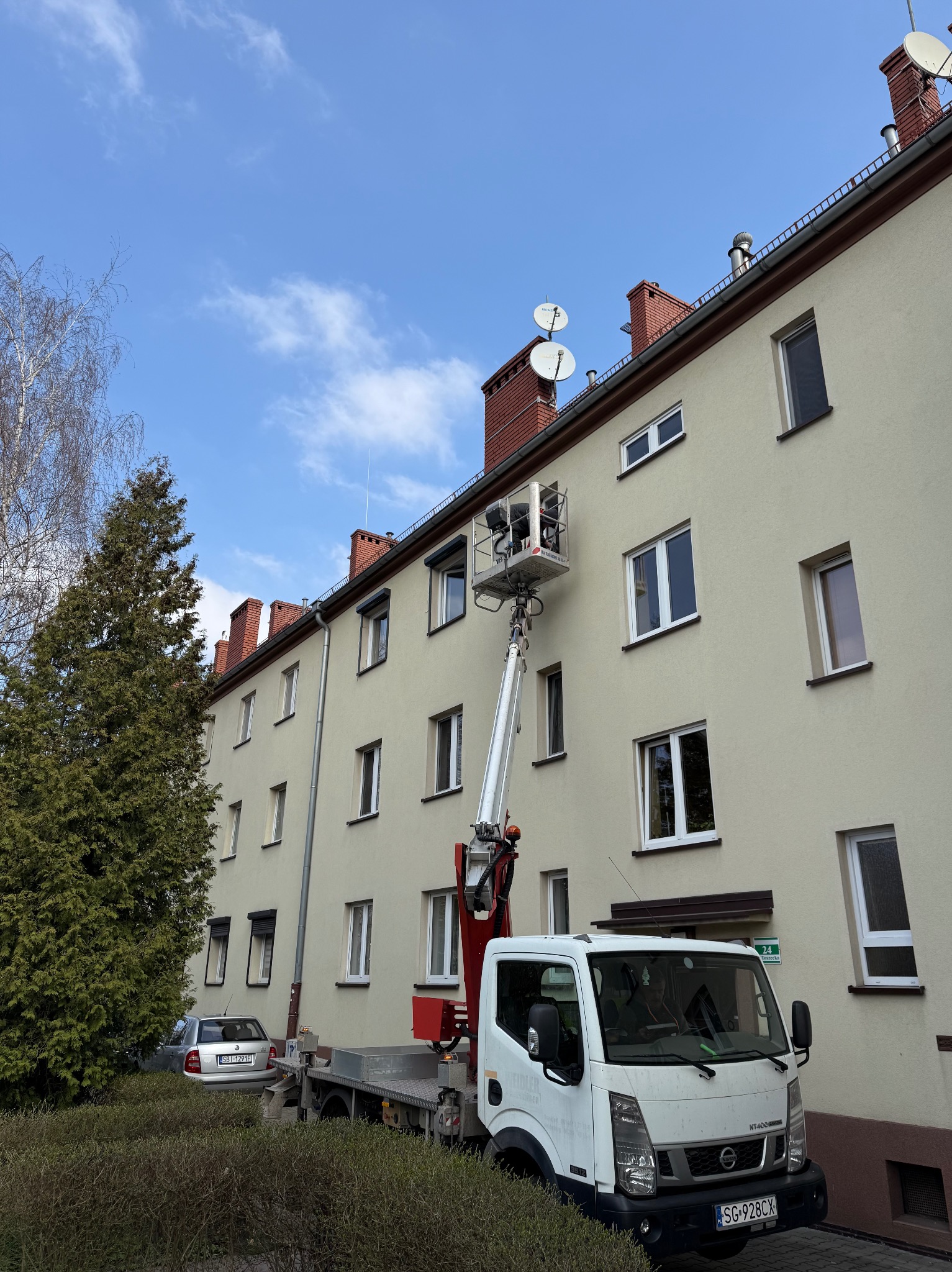 Montażysta w podnośniku koszowym instaluje osłonę okienną na elewacji budynku mieszkalnego, widoczny samochód dostawczy z podnośnikiem i fragment zieleni.