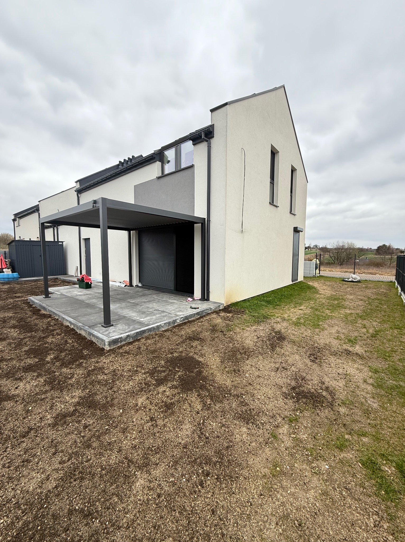 Nowoczesna, szara pergola metalowa z roletą zewnętrzną przy białym domu z kostką brukową na patio, widok z perspektywy ogrodu w pochmurny dzień.