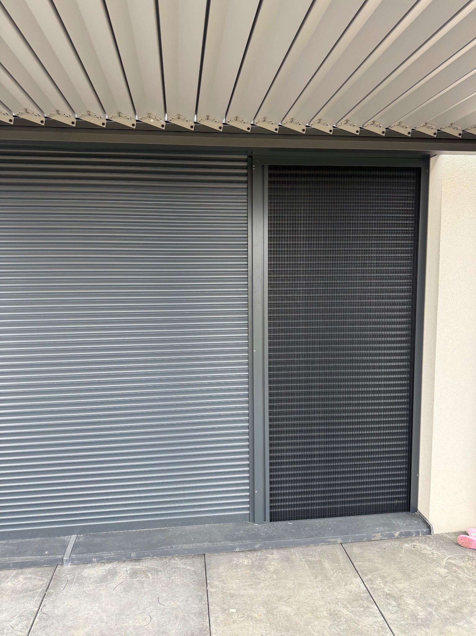 Zewnętrzna szara roleta obok czarnej moskitiery ramkowej, zamontowane pod jasną metalową konstrukcją zadaszenia.