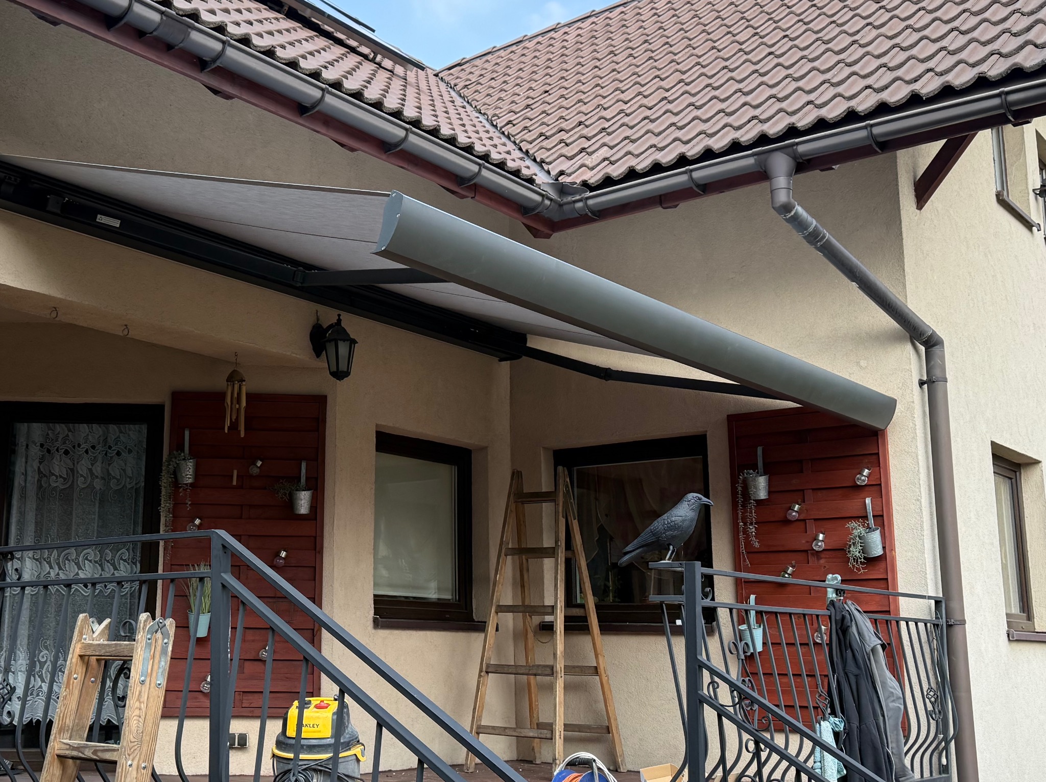 Szara, zwinięta markiza tarasowa zamontowana nad balkonem domu z brązową dachówką, widoczna drabina, dekoracje balkonowe i atrapa kruka na balustradzie.