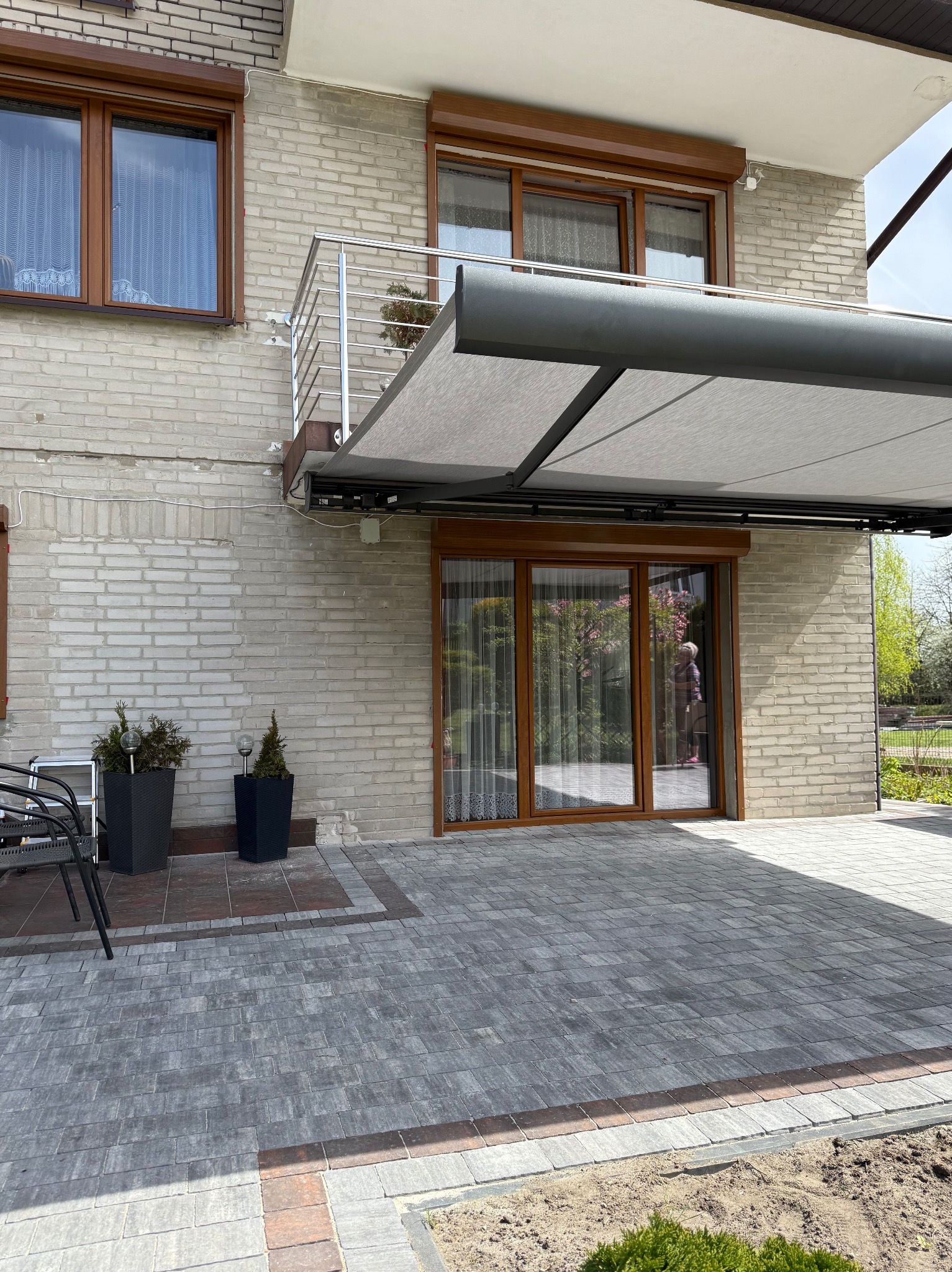 Zewnętrzna pergola metalowa z jasną tkaniną zamontowana nad tarasem przy domu z jasnej cegły, widoczne okna z roletami zewnętrznymi w brązowych ramach oraz szara kostka brukowa.
