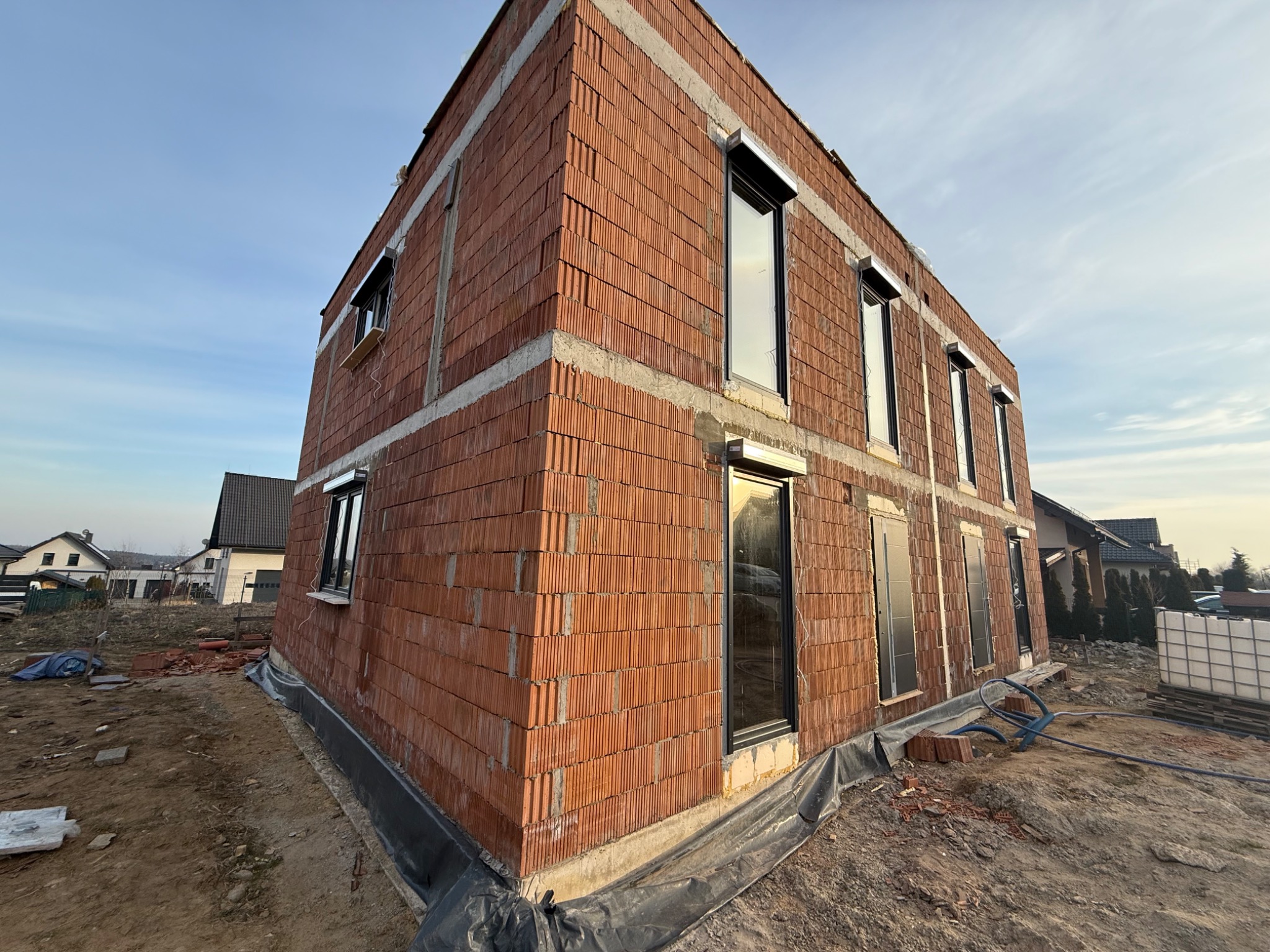 Surowy budynek z czerwonej cegły z zamontowanymi oknami i przygotowanymi otworami na rolety zewnętrzne, widok z zewnątrz na placu budowy.