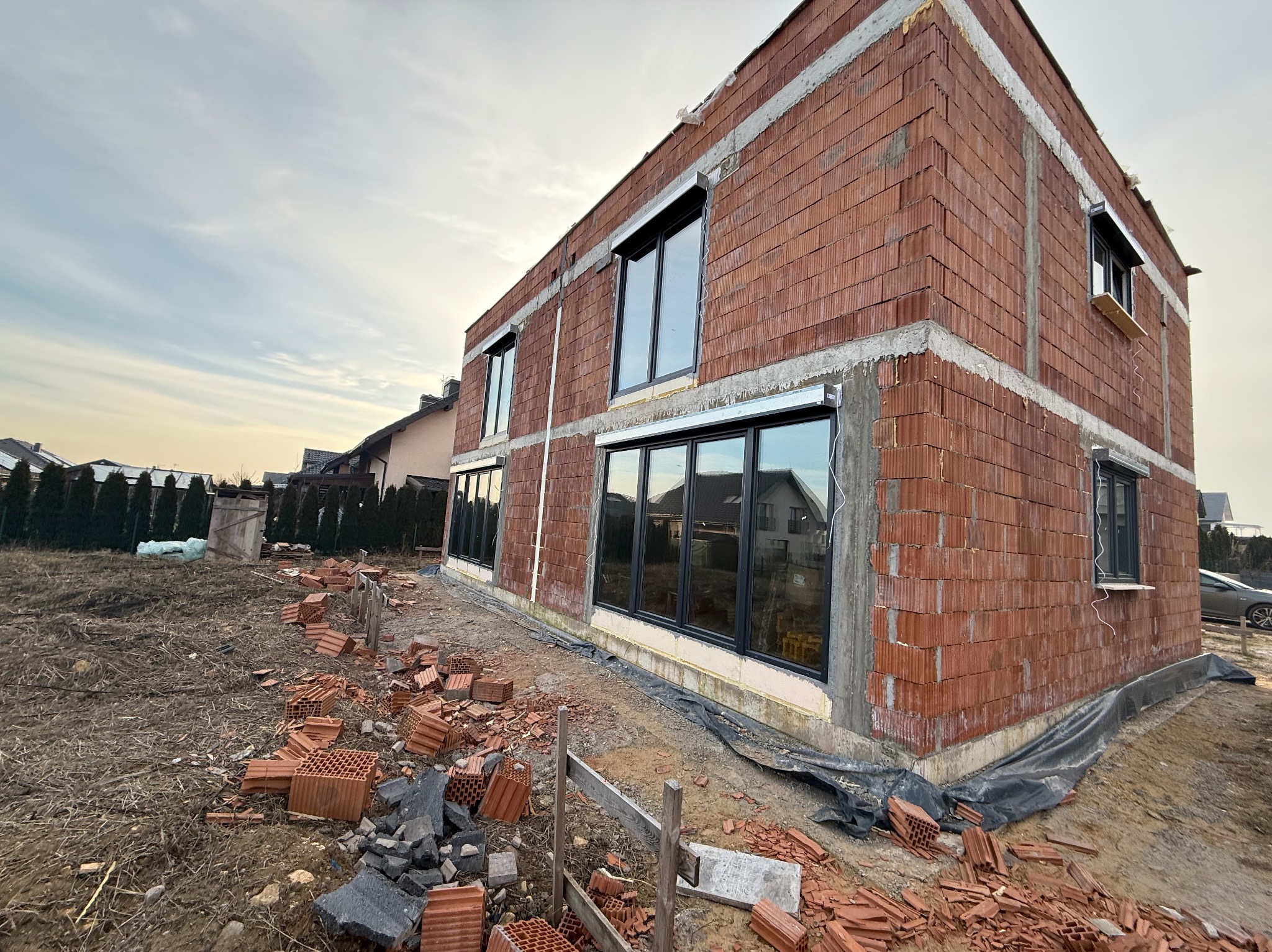 Surowy stan budynku z zamontowanymi oknami i przygotowanymi otworami na rolety zewnętrzne, widoczne porozrzucane cegły na posesji.
