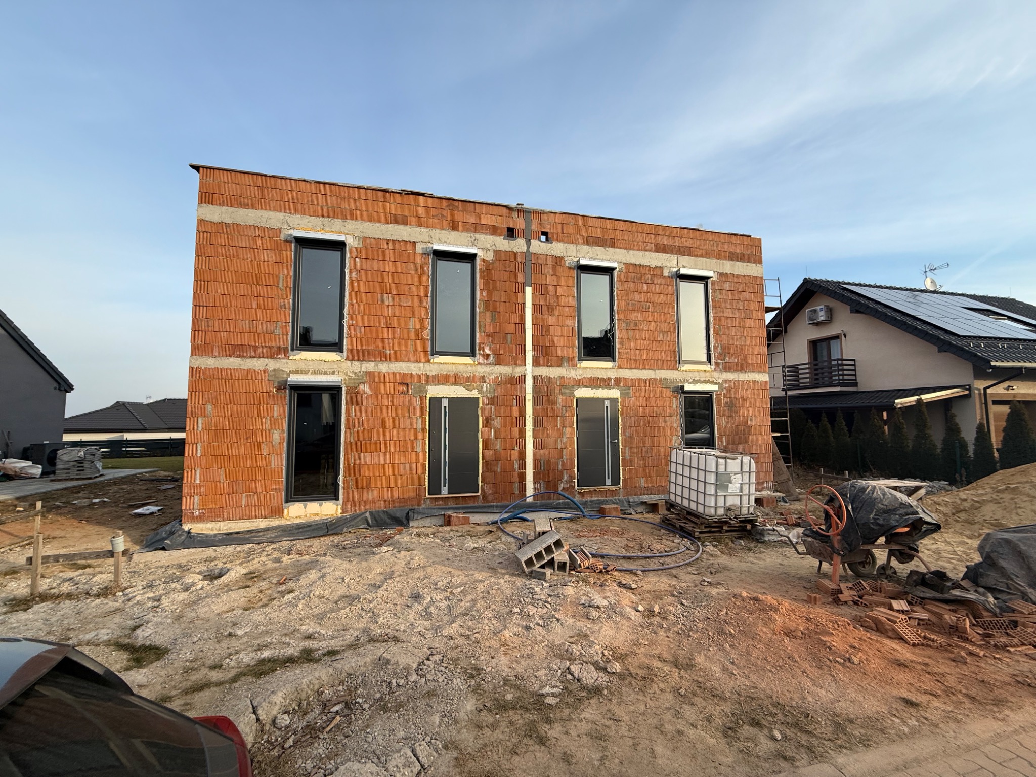 Surowy stan budynku z czerwonej cegły z zamontowanymi oknami i drzwiami, widoczna betoniarka i materiały budowlane na placu budowy.