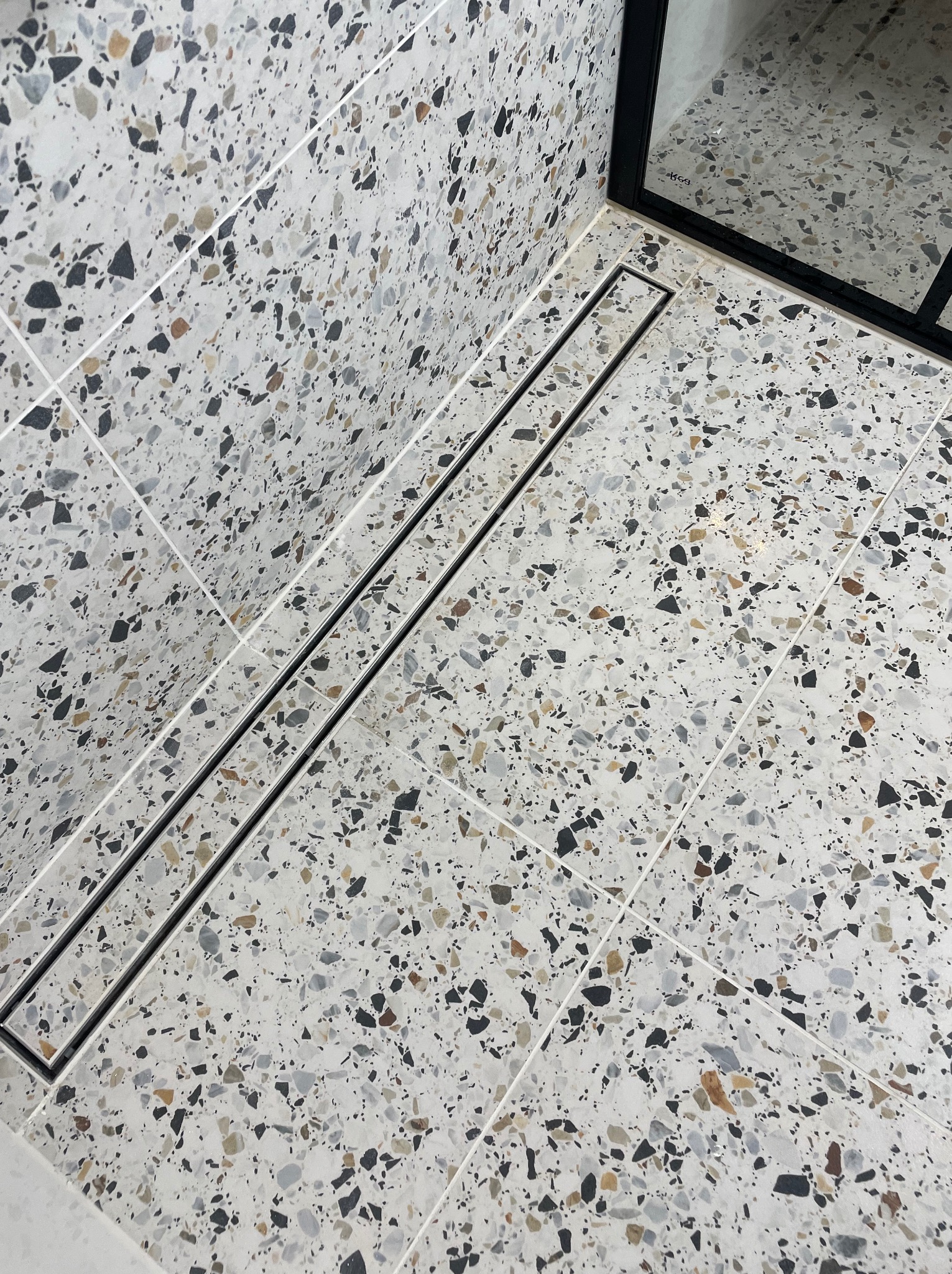 Nowoczesna łazienka z podłogą i ścianami wyłożonymi płytkami imitującymi lastryko, z czarnym odpływem liniowym i fragmentem kabiny prysznicowej z czarną ramą.