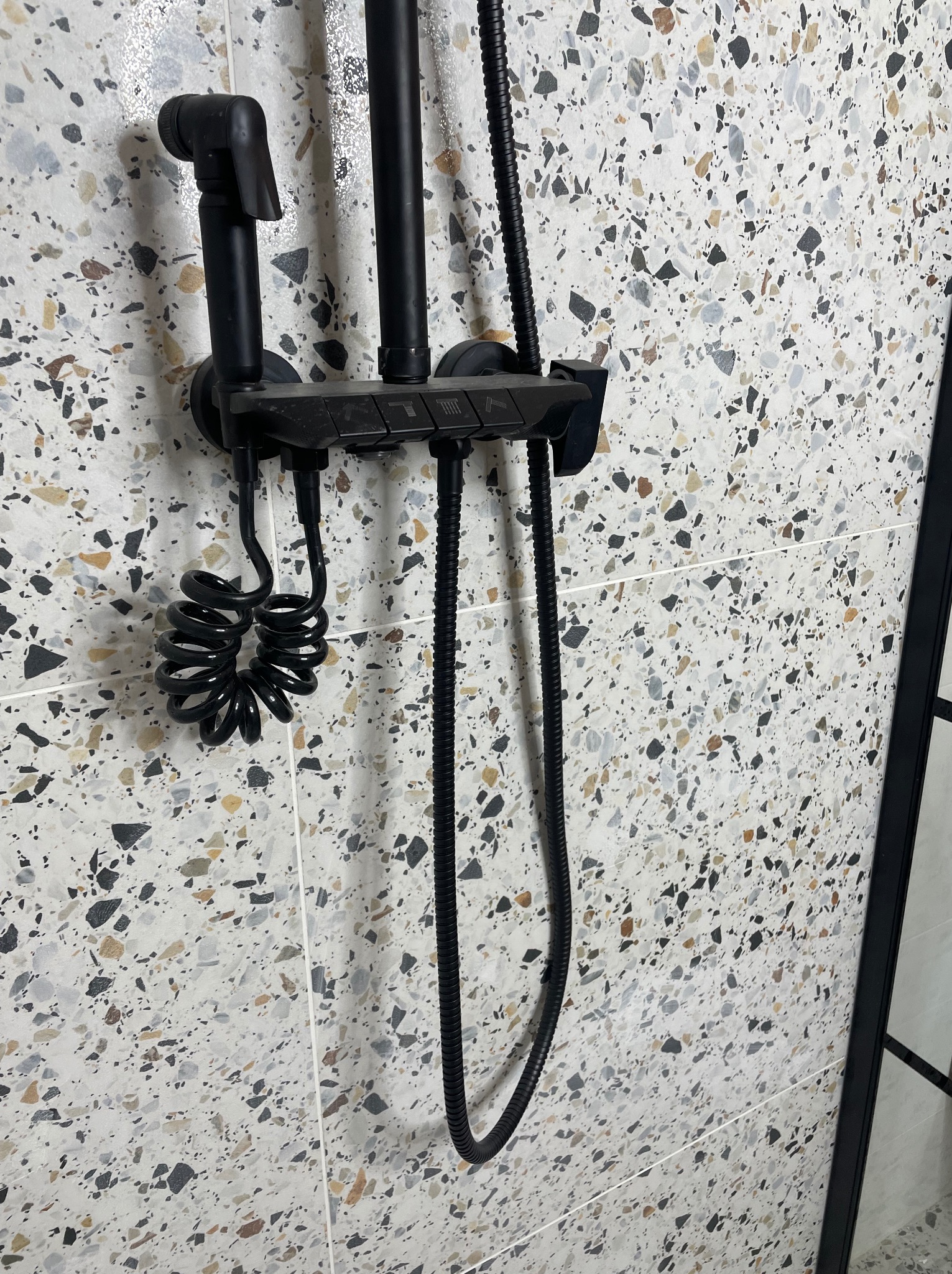 Czarny zestaw natryskowy z bidetką na ścianie wyłożonej płytkami imitującymi lastryko w łazience.