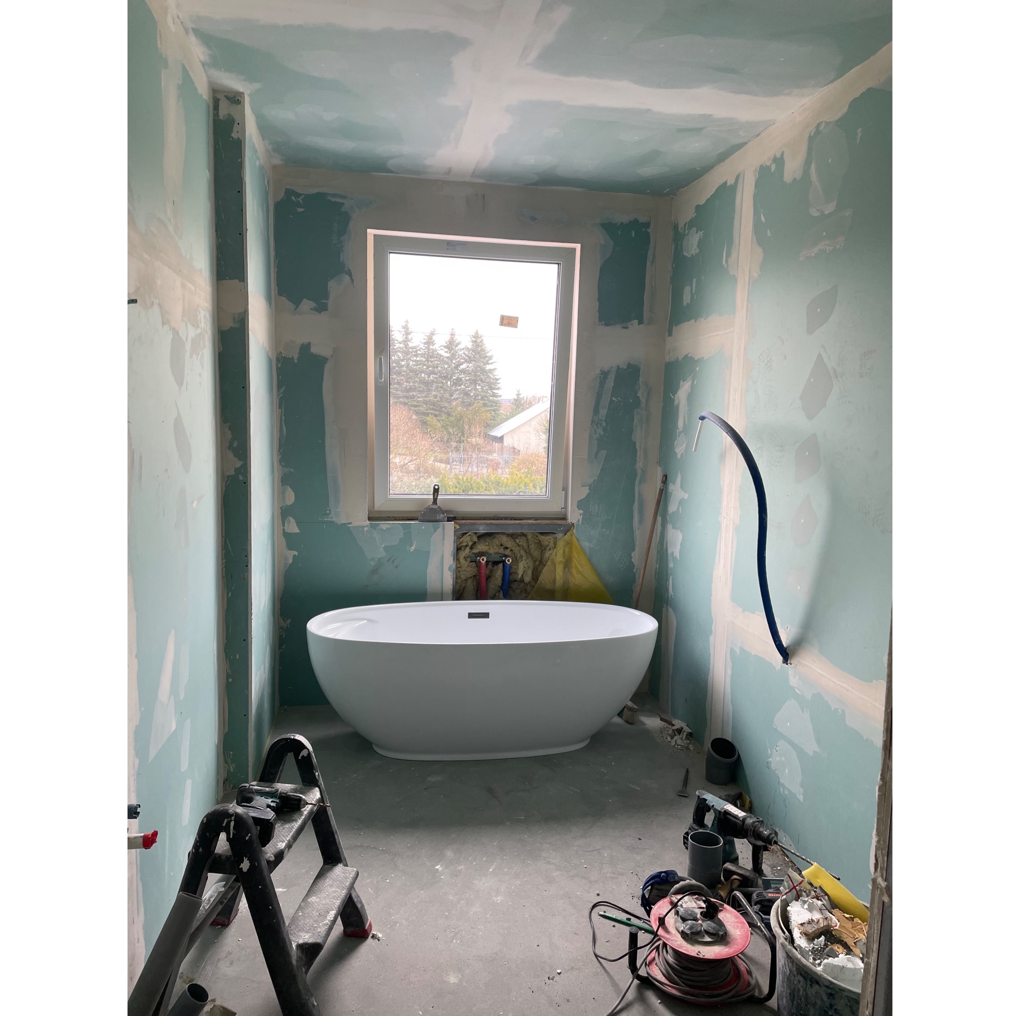 Łazienka w trakcie prac wykończeniowych: owalna wanna wolnostojąca, ściany przygotowane pod gładzie, widoczne narzędzia i drabina, okno z widokiem na dom i drzewa.