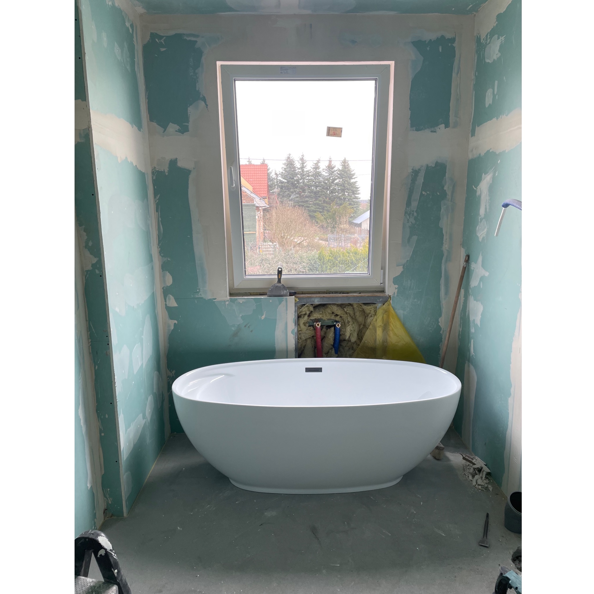 Wolnostojąca biała wanna owalna w surowym wnętrzu łazienki, ściany z płyt gipsowo-kartonowych przed wykończeniem, widoczne instalacje wodne, szpachelka na parapecie okna z widokiem na drzewa...