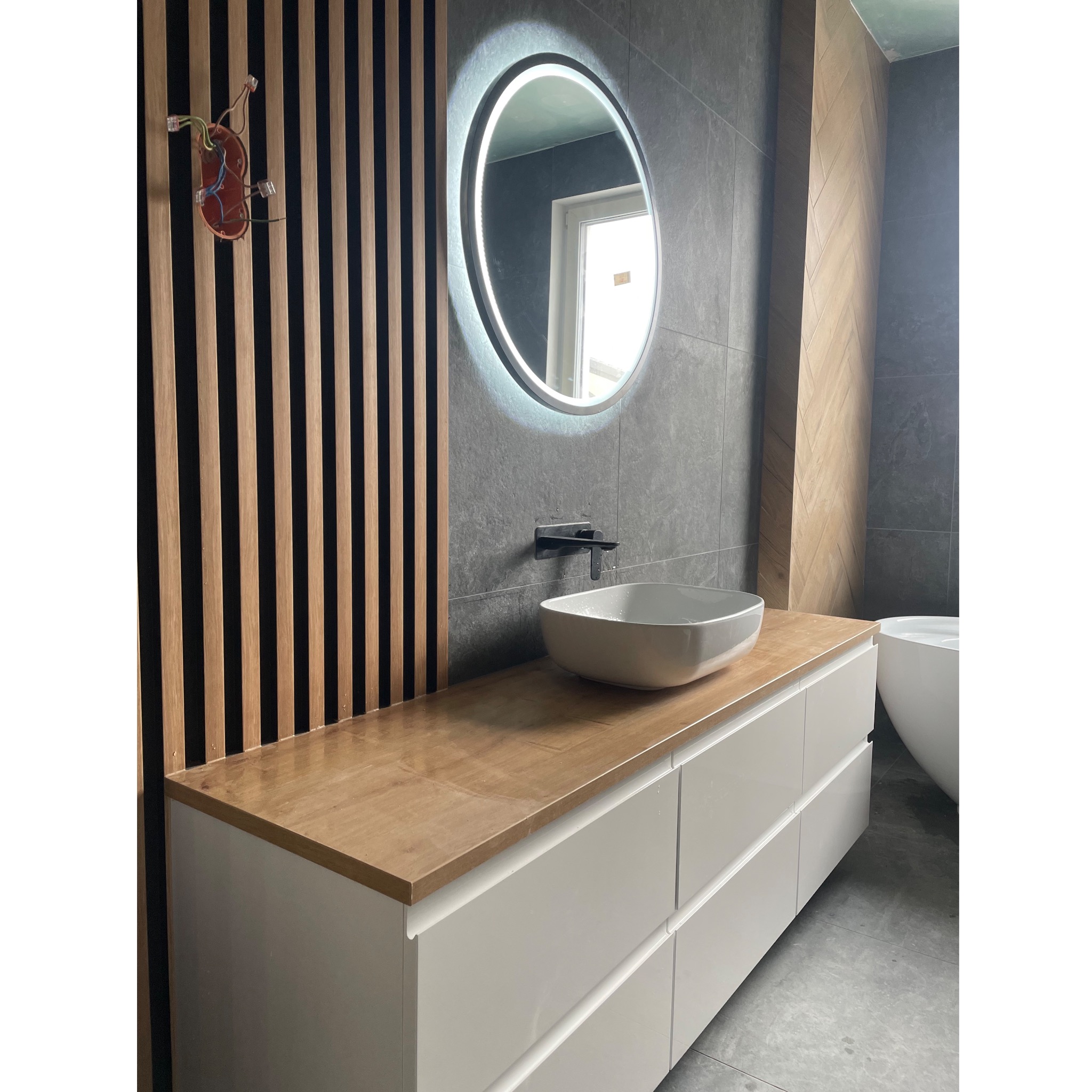 Nowoczesna łazienka z umywalką nablatową, podświetlanym lustrem i drewnianymi lamelami na ścianie. Widoczne przyłącza elektryczne na ścianie.