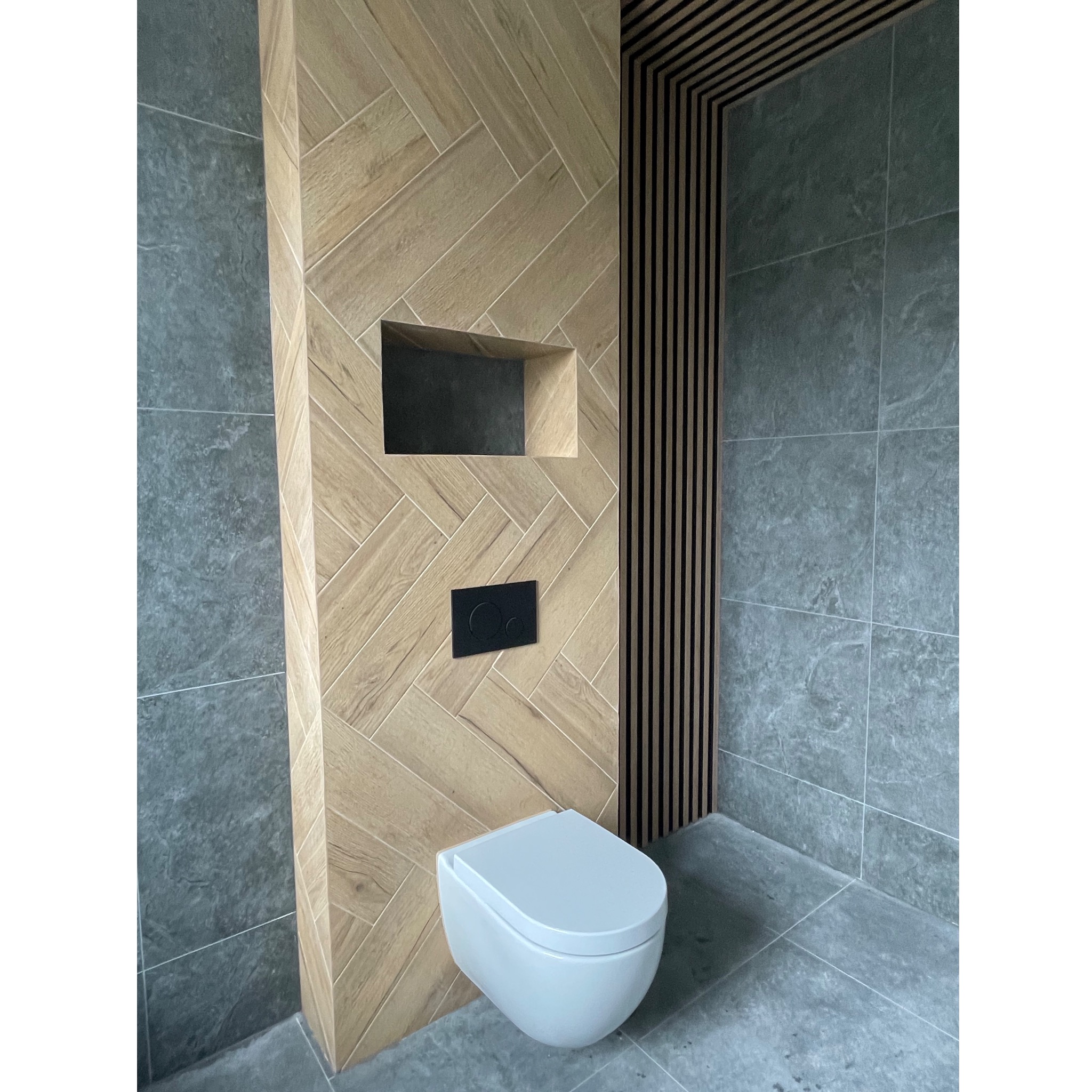 Nowoczesna łazienka z wiszącą toaletą, drewnopodobnymi płytkami ułożonymi w jodełkę na ścianie, czarnym przyciskiem spłukującym i pionowymi drewnianymi listwami dekoracyjnymi.