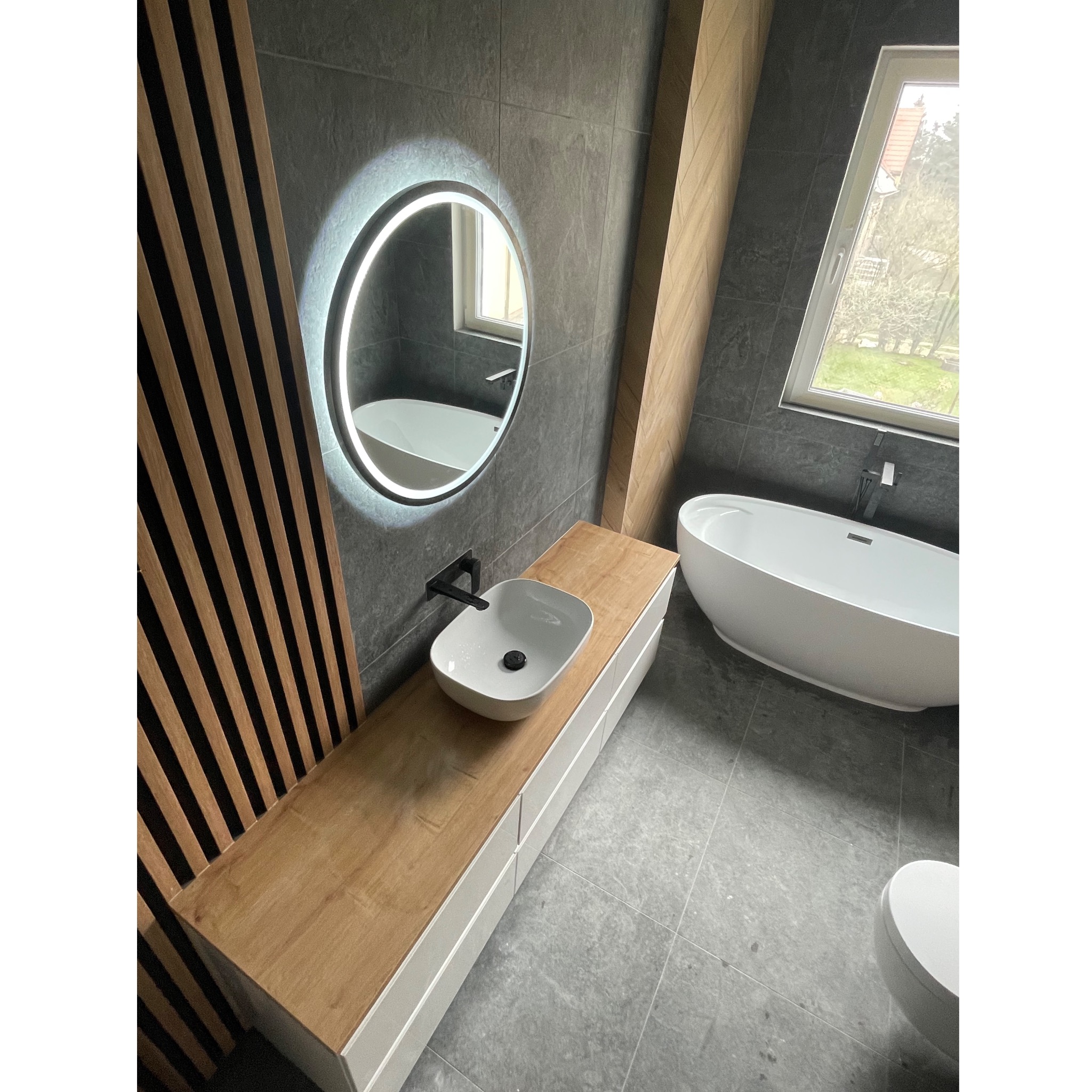 Nowoczesna łazienka z szarymi płytkami, drewnianymi panelami dekoracyjnymi, okrągłym lustrem z podświetleniem LED, białą umywalką nablatową z czarną armaturą i wolnostojącą wanną.