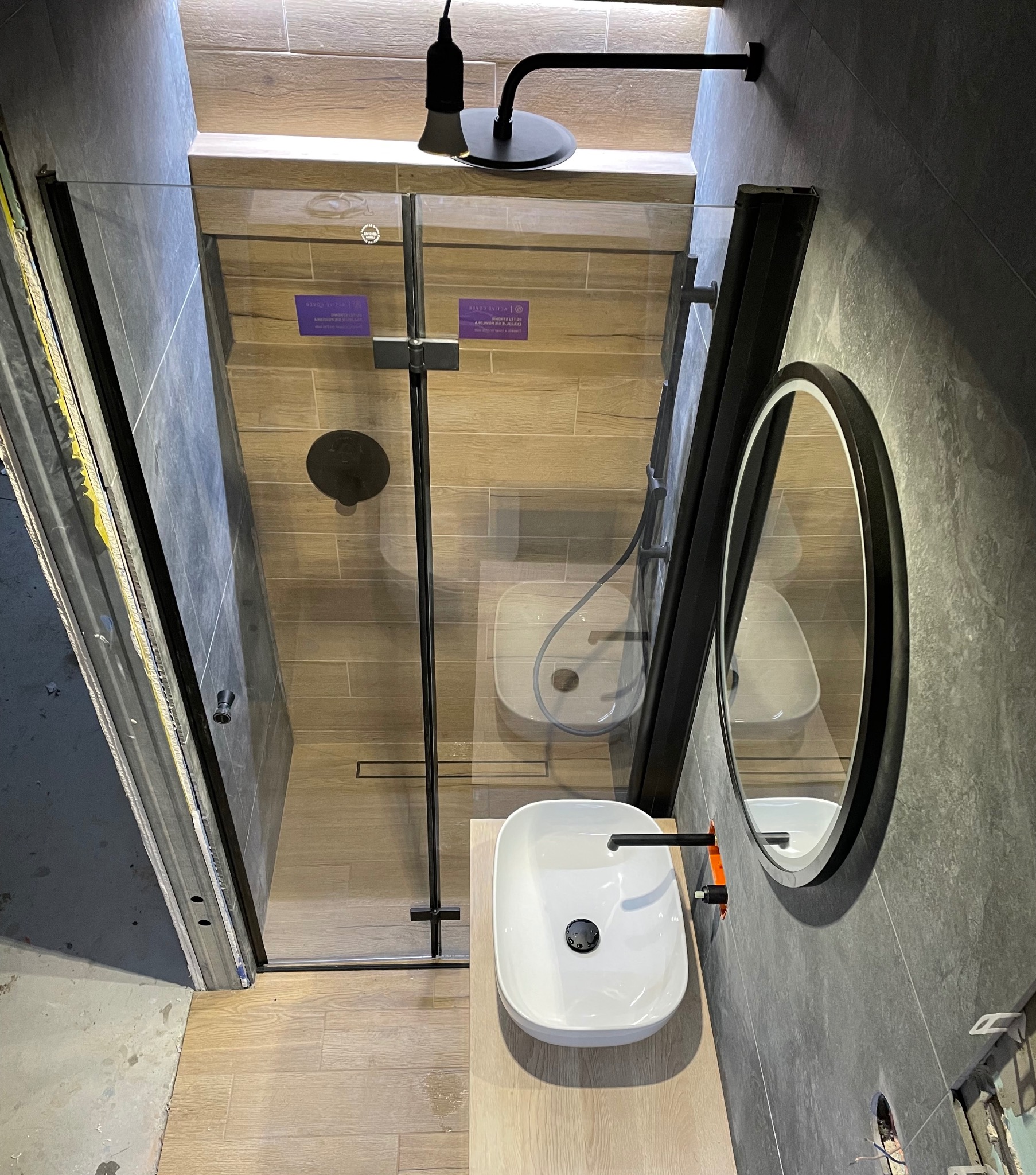 Nowoczesna łazienka z kabiną prysznicową typu walk-in z czarnymi profilami, owalnym lustrem w czarnej ramie i białą umywalką nablatową z czarną armaturą, widok z góry, fragment niedokończonej...