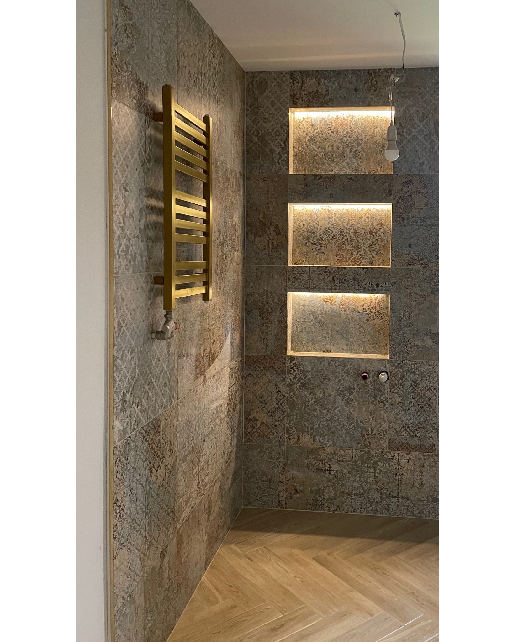 Wykończona łazienka z dekoracyjnymi płytkami ściennymi, złotym grzejnikiem drabinkowym i podświetlanymi wnękami w ścianie. Na podłodze parkiet ułożony we wzór jodełki.